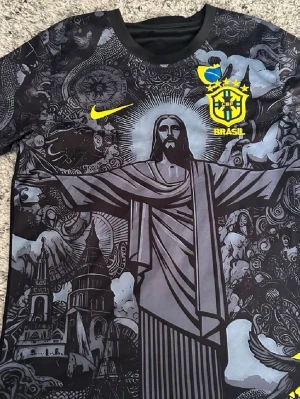 Svart Brasilien Nike fotbollströja - Unik svart fotbollströja från Nike med Brasilien-emblem och gult Nike-märke. Framsidan har ett stort tryck av Kristusstatyn och detaljerade grafiska mönster i grått. Baksidan är helsvart med Brasilien-logga upptill. Perfekt för dig som älskar snygga matchtröjor.