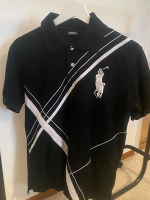 Svart piké från Polo Ralph Lauren - Svart pikétröja från Polo Ralph Lauren med vita diagonala ränder och stor broderad logga på bröstet. Klassisk krage, knappar framtill och korta ärmar. Siffran 3 i vitt på ena ärmen ger en sportig touch. Perfekt för en stilren och avslappnad look.
