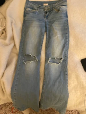 Ljusblå slitna jeans från Nelly - Säljer ett par ljusblå jeans från Nelly med utsvängda ben och stora slitningar över knäna. Klassisk femficksmodell i mjukt denimtyg. Perfekta för dig som gillar en avslappnad och trendig stil. Några defekter den vänstra knappen har lossnat och hål där under men absolut inget som syns !