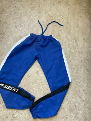 Blåa mjukisbyxor från Lacoste - Snygga blåa mjukisbyxor från Lacoste med vita sidoremsor och svart detalj med logga på ena benet. Byxorna har resår i midjan med dragsko och muddar vid bensluten. Perfekta för en avslappnad och sportig stil.