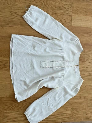 Vit blus med dekorativ knappslå - Stilren vit blus från mayla med långärmad design och rund halsringning. Framtill finns en dekorativ knappslå med små tygklädda knappar. Blusen har lätt puffade axlar och ärmar med resår vid ärmslut. Tillverkad i ett mjukt, vävt material.