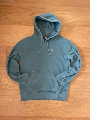 Blågrön hoodie från Dickies - Snygg blågrön hoodie från Dickies med klassisk känguruficka och liten Dickies-logga på bröstet. Tröjan har huva med dragsko och ribbade muddar vid ärmslut och nederkant. Perfekt för en avslappnad och stilren look.