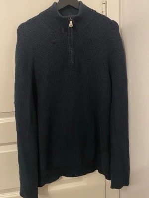 Mörkblå stickad tröja Henri Lloyd - Snygg mörkblå stickad tröja från Henri Lloyd med halv dragkedja och hög krage. Tröjan har grovstickad struktur och ribbade muddar vid ärmslut och nederkant. Perfekt för kyliga dagar och enkel att matcha med jeans eller chinos.