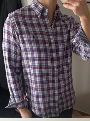 Rutig skjorta från Ralph Lauren - Klassisk rutig skjorta från Ralph Lauren i blått, rött och vitt. Skjortan har lång ärm, knappar framtill och button-down krage. Tillverkad i mjuk bomull och har den ikoniska broderade loggan på bröstet. Perfekt för en avslappnad men ändå stilren look.