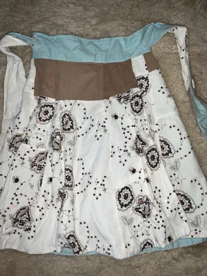 Vit omlottkjol med blommigt mönster - Unik omlottkjol i vitt med blommigt mönster och broderade detaljer. Kjolen har bred midja i beige och ljusblått tyg samt knytband i midjan. Perfekt för dig som gillar boho-stil och vill ha något annorlunda.