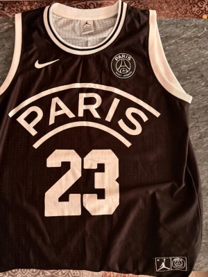 Jordan x PSG svart basketlinne - Snyggt svart basketlinne från Jordan x Paris Saint-Germain med vita detaljer, stort tryck med 'PARIS' och nummer 23 fram samt 'JORDAN' och 23 bak. Tillverkat i meshmaterial som andas, perfekt för träning eller streetwear.