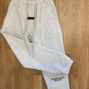  Oatmeal Grey Essentials byxor - Säljer ett par ljusgrå mjukisbyxor från Fear of God Essentials. Byxorna har resår i midjan med långa dragsnören, muddar vid bensluten och logga tryckt på benet. Perfekta för en avslappnad och trendig look. Obs! Priset är ej hugget i sten!