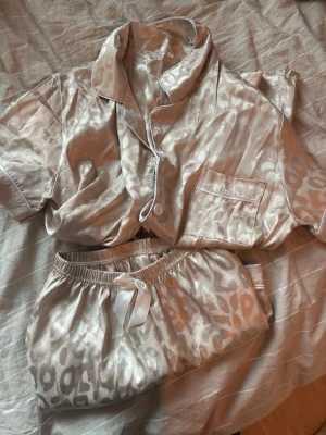Beige satinpyjamas med leopardmönster - Snygg pyjamas i beige satin med subtilt leopardmönster. Setet består av en kortärmad skjorta med vit piping och bröstficka samt matchande shorts med elastisk midja och vit rosett. Perfekt för en lyxig och avslappnad kväll. Storlek S men passar M 