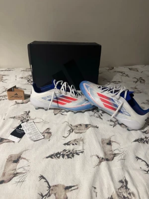Elite Adidas F50 vita/blå/röda fotbollsskor - Säljer ett par Elite Adidas F50 fotbollsskor i vitt med blå och röda detaljer. Skorna har klassisk låg profil, snörning och mönstrad yttersula för bra grepp på planen. Tillverkade i syntetmaterial med ventilerande mesh och ikoniska Adidas-ränder på sidan. Använda en gång bara, testat dem. 