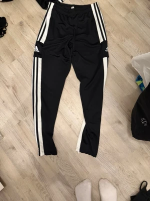 Svarta Adidas mjukisbyxor med vita ränder - Säljer ett par svarta mjukisbyxor från Adidas med klassiska vita ränder längs benen och dragkedjor vid bensluten. Byxorna har resår i midjan och är tillverkade i ett mjukt, stretchigt material som passar perfekt för chill eller träning.