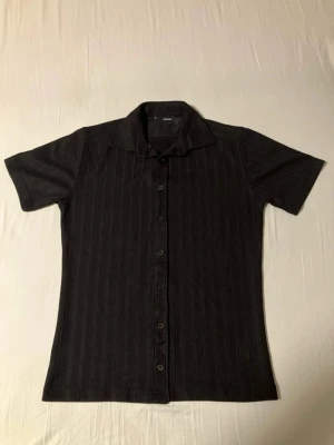 Svart kortärmad skjorta med knappar - Stilren svart skjorta med diskreta vertikala ränder och klassisk krage. Skjortan har korta ärmar och knäppning framtill. Perfekt för en clean och enkel look. Materialet känns mjukt och bekvämt, troligen bomull eller polyester.