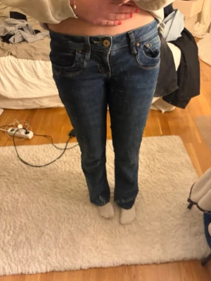 Mörkblå bootcut jeans med låg midja - Säljer ett par mörkblå bootcut jeans med låg midja och klassiska fem fickor. Jeansen har guldfärgade sömmar och knappdetalj framtill. Materialet är denim i bomull och passformen är figurnära upptill med utsvängda ben.