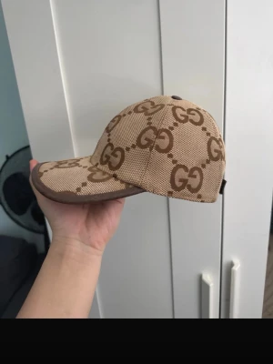 Jumbo GUCCI keps - Säljer en beige keps från Gucci med det klassiska GG-monogrammet i brunt över hela kepsen. Kepsen har böjd skärm, justerbart band bak och är tillverkad i canvasmaterial med bruna detaljer. Perfekt för dig som vill ha en stilren och lyxig accessoar.