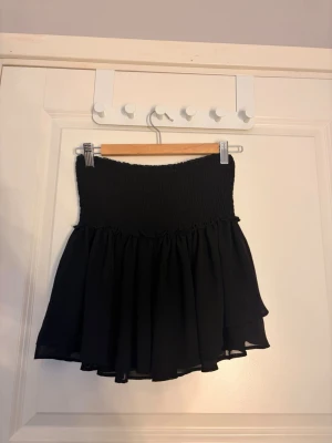 Svart volangkjol - Supersöt svart kortkjol med smockad midja och volangig nederdel. Kjolen har en flowy passform och är perfekt för dig som gillar en enkel men ändå stylish look. Tillverkad i ett lätt och luftigt material.