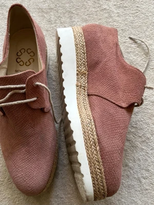 Rosa mocka loafers från Sapatos - Säljer ett par snygga rosa loafers från Sapatos i mocka med struktur. Skorna har vita snören och en platåsula med flätad detalj i naturfärg samt vit räfflad undersida. Perfekta för dig som vill ha något unikt och trendigt till din outfit.