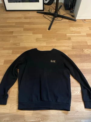 Svart sweatshirt från EA7 - Snygg svart sweatshirt från EA7 med rund halsringning och diskret logga i guld på bröstet. Tröjan har långa ärmar och ribbade muddar vid ärmslut och nederkant. Perfekt för en clean och stilren look.