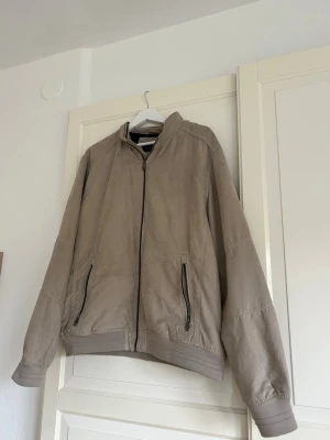 Beige mockajacka med dragkedja - Beige mockajacka med hel dragkedja framtill och två fickor med svarta dragkedjor!! Två defekter på jackan. 