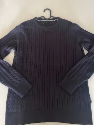 Mörkblå kabelstickad tröja  - Säljer en mörkblå kabelstickad tröja från Lindbergh med rund halsringning och långa ärmar. Tröjan har ett klassiskt kabelmönster och ribbade muddar vid ärmslut och nederkant. Perfekt för dig som gillar stilrena och tidlösa plagg. Helt ny säljer för att den aldrig användes. Nypris 799kr, mitt pris 399kr. 