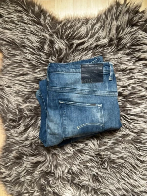 Blå jeans från G-Star RAW  - Säljer ett par klassiska blå jeans från G-Star RAW med slitningar och tvättade detaljer. Jeansen har raka ben, fem fickor och en mörk läderpatch med logga bak i midjan. Perfekta för en avslappnad och trendig look.