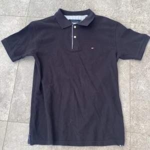 Svart pikétröja från Tommy Hilfiger - Klassisk svart pikétröja från Tommy Hilfiger med korta ärmar och tre knappar framtill. Tröjan har en liten broderad logga på bröstet och är gjord i bomull. Snygg krage och diskreta randiga detaljer på insidan av kragen. 9/10 skick, hör av er vid frågor✅