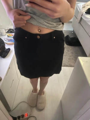 Svart kortkjol med fickor - Säljer en svart kortkjol med två framfickor och knappdetalj i midjan. Kjolen har en enkel och stilren design som passar till många olika outfits. Perfekt för dig som gillar en klassisk look.