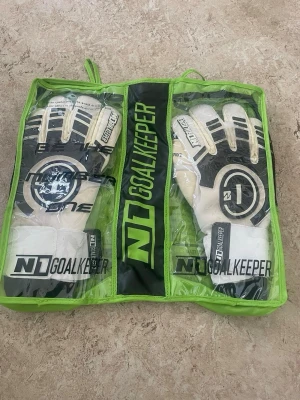 N1 Goalkeeper målvaktshandskar - Snygga målvaktshandskar från N1 Goalkeeper i vitt och svart med coola detaljer och breda muddar. Perfekta för dig som vill ta ditt spel till nästa nivå. Handskarna har en sportig look och kommer i en praktisk grön förvaringsväska.