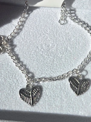 Silverfärgat armband med hjärtberlocker Rostfritt Stål  - Stilrent armband i silverfärgat rostfritt stål med flera hjärtformade berlocker dekorerade med vingar. Kedjan är tunn och har justerbar längd med karbinlås. Perfekt accessoar för dig som gillar detaljer med symbolik och en unik touch. Handgjort.