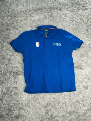 Blå pikétröja från BOSS  - Snygg blå pikétröja från BOSS med korta ärmar och klassisk krage. På bröstet finns ett broderat BOSS New York-tryck och en sköld i italienska flaggans färger. Kragen har också en detalj i grönt, vitt och rött på baksidan.
