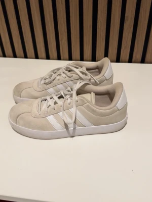 Beige Adidas sneakers med mockadetaljer - Snygga beige sneakers från Adidas med klassiska vita ränder på sidorna. Skorna har en låg profil, rund tå och ovandel i mocka och textil. Tjock vit sula och vita skosnören ger en clean look som passar till det mesta. 