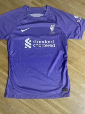Lila Liverpool FC matchtröja Nike - Snygg lila matchtröja från Liverpool FC med Nike-logga och klubbmärke på bröstet. Tröjan har korta ärmar och tryckt sponsorlogga framtill. Perfekt för dig som älskar fotboll och vill visa ditt stöd för LFC på planen eller läktaren.
