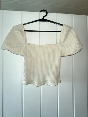 Vit ribbad topp med puffärm - Trendig vit ribbad topp med fyrkantig urringning och stora puffärmar. Croppad modell som ger en snygg siluett och passar perfekt till både jeans och kjol. Materialet är mjukt och stretchigt för extra komfort.