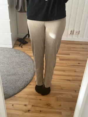 Beige ribbade mjukisbyxor - Säljer ett par beige mjukisbyxor med smala vita ränder och ribbad struktur. Byxorna har rak passform och är stretchiga för extra komfort. Perfekta för chill dagar hemma eller när du vill ha det bekvämt. Materialet känns mjukt och följsamt.