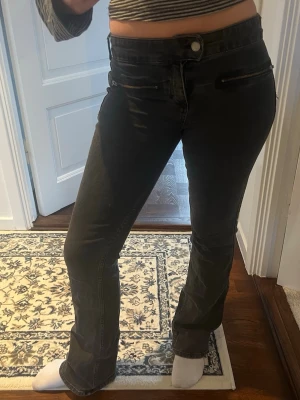 Svarta bootcut jeans med dragkedjor - Snygga svarta bootcut jeans med två dekorativa dragkedjor framtill. Jeansen har låg midja och raka ben med lätt utsvängd passform. Perfekta för en trendig och avslappnad stil. Ifrån pull&bear strl 38. Lite slitna nertill vid foten.