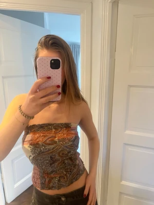 Mönstrad bandeau tubtopp i paisley - Säljer en snygg bandeau tubtopp med paisleyinspirerat mönster i orange, brun, beige och svart. Toppen är axelbandslös och har en croppad passform som sitter tight mot kroppen. Perfekt för sommardagar och festivaler! Strl S ifrån next petite