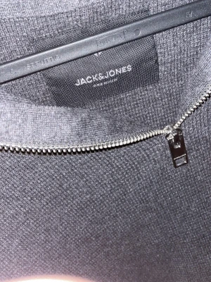 Mörkgrå halvzip från Jack & Jones - Mörkgrå stickad tröja från Jack & Jones med halv dragkedja framtill. Tröjan har hög krage och är tillverkad i mjukt material, perfekt för lager på lager. Enkel och stilren design som passar till det mesta.