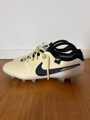 Nike Tiempo Legend 8 elite FG fotbollsskor - Snygga fotbollsskor från Nike, modell Tiempo Legend 8 Pro FG, i beige och svart med gulddetaljer. Skorna har klassisk snörning, struktur i lädret för bättre bollkontroll och en stabil yttersula med fasta dobbar för spel på naturgräs. Perfekta för dig som vill ha stil och prestanda på planen.