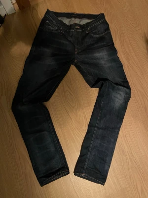 Mörkblå jeans från Nudie Jeans - Säljer ett par mörkblå jeans från Nudie Jeans med klassisk femficksmodell och kontrastsömmar. Jeansen har en smal passform och snygga slitningar framtill. Bakfickorna har Nudie Jeans signaturbroderi. Perfekta för en avslappnad och stilren look.