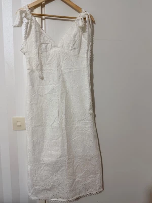 Vit spetsklänning med knytaxlar - Söt vit klänning i bomull med broderad spets och knytbara axelband. Klänningen har v-ringning och är vadlång med fina spetsdetaljer längs kanterna. Perfekt för sommarens alla tillfällen och ger en romantisk vibe.