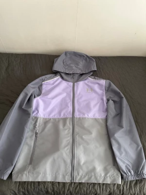Under Armour Vindjacka  - Snygg vindjacka från Under Armour i grått och lila. Jackan har huva, hel dragkedja framtill och två sidofickor med dragkedja. Tillverkad i lätt och vindtätt material, perfekt för blåsiga dagar. Under Armour-logga på bröstet.  Storlek XS 