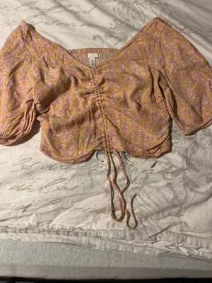 Blommig omlottopp med snörning - Supersöt blommig topp från H&M i ljusgult och lila med omlottkänsla och snörning framtill. Toppen har korta puffärmar och croppad passform, perfekt för varma dagar. Materialet känns lätt och luftigt.