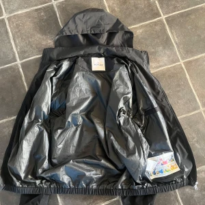 Svart vindjacka från Moncler - Snygg svart vindjacka från Moncler med huva och elastiska muddar. Jackan har en ficka på ärmen med Moncler-logga och innerfoder i blankt material. Perfekt för blåsiga dagar och enkel att matcha med olika outfits. Pris ej hugget i sten, fraktar inom 1-2 dagar