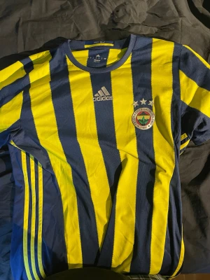 Vintage Fenerbahce fotbollströja - Säljer en Fenerbahce matchtröja från Adidas i gul och marinblå med vertikala ränder. Tröjan har klubbmärke och Adidas-logga på bröstet samt tre stjärnor ovanför klubbmärket. Tillverkad i lätt och ventilerande material, perfekt för fotboll eller träning.