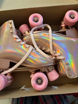 Holografiska rullskridskor med rosa hjul - Säljer ett par riktigt snygga rullskridskor i holografiskt material med högt skaft och rosa snörning. De har stabila rosa hjul och tåstopp, perfekta för dig som vill sticka ut på rinken eller gatan. Skorna har en cool glansig finish som skiftar i regnbågens färger.
