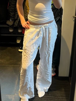 Vita linnebyxor med snören - Säljer ett par vita linnebyxor med avslappnad passform och dragsko i midjan. Byxorna har raka ben och elastisk midja för extra komfort. Perfekta för en chill och stilren look. Materialet är lätt och luftigt linne.
