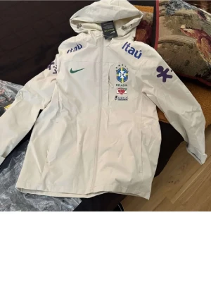 Vit Brasil Nike vindjacka med huva - Snygg vit vindjacka från Nike med Brasiliens landslagslogga och sponsortryck i blått och lila. Jackan har hel dragkedja, två sidofickor och huva. Perfekt för dig som gillar sportig stil och vill sticka ut med landslagsvibbar.