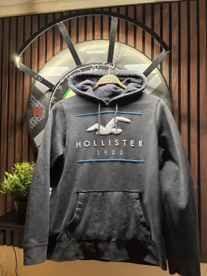  Hollister hoodie - Snygg mörkgrå hoodie från Hollister med stor logga och texten 'Hollister 1922' broderat på bröstet. Hoodien har en klassisk känguruficka, dragsko i huvan och blå detaljer. Perfekt för en chill och avslappnad stil.