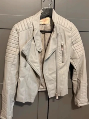 Ljusgrå biker skinnjacka - Snygg ljusgrå skinnjacka i bikerstil med dragkedja snett framtill och quiltade detaljer på axlar och ärmar. Jackan har en bröstficka med dragkedja och markerad krage. Storlek M , liten i storleken . Fint skick . 