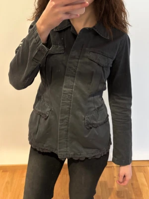 Svart safarijacka från Odd Molly - Snygg svart rock med klassisk krage och flera fickor framtill. Jackan har broderad text på ena ärmen och stängs med knappar. Materialet är bomull och modellen har en normal passform med långa ärmar. Storlek M men passar mer xs/s!!