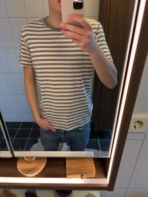 Randig t-shirt med beige och vitt - Säljer en klassisk randig t-shirt i beige och vitt med rund halsringning och korta ärmar. T-shirten har en relaxed passform och är tillverkad i mjuk bomull, perfekt för en chill och enkel stil.
