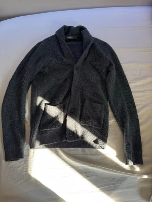 Mörkgrå stickad kofta med fickor - Mörkgrå stickad kofta med knappar framtill och två stora fickor. Koftan har en bred krage och är ribbstickad för extra struktur. Perfekt för en clean stil.